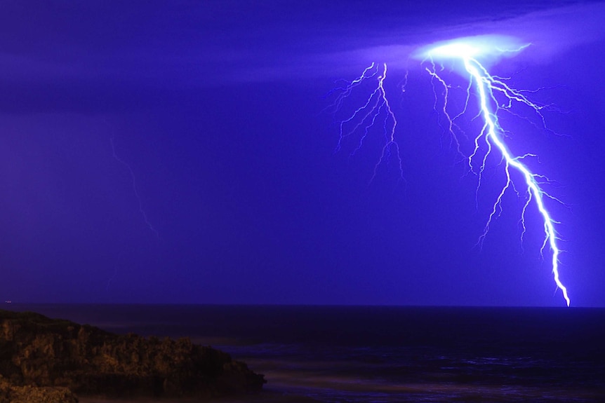 A massive bolt of lightning cuts through a blue sky, with part of the coast lit up by it.