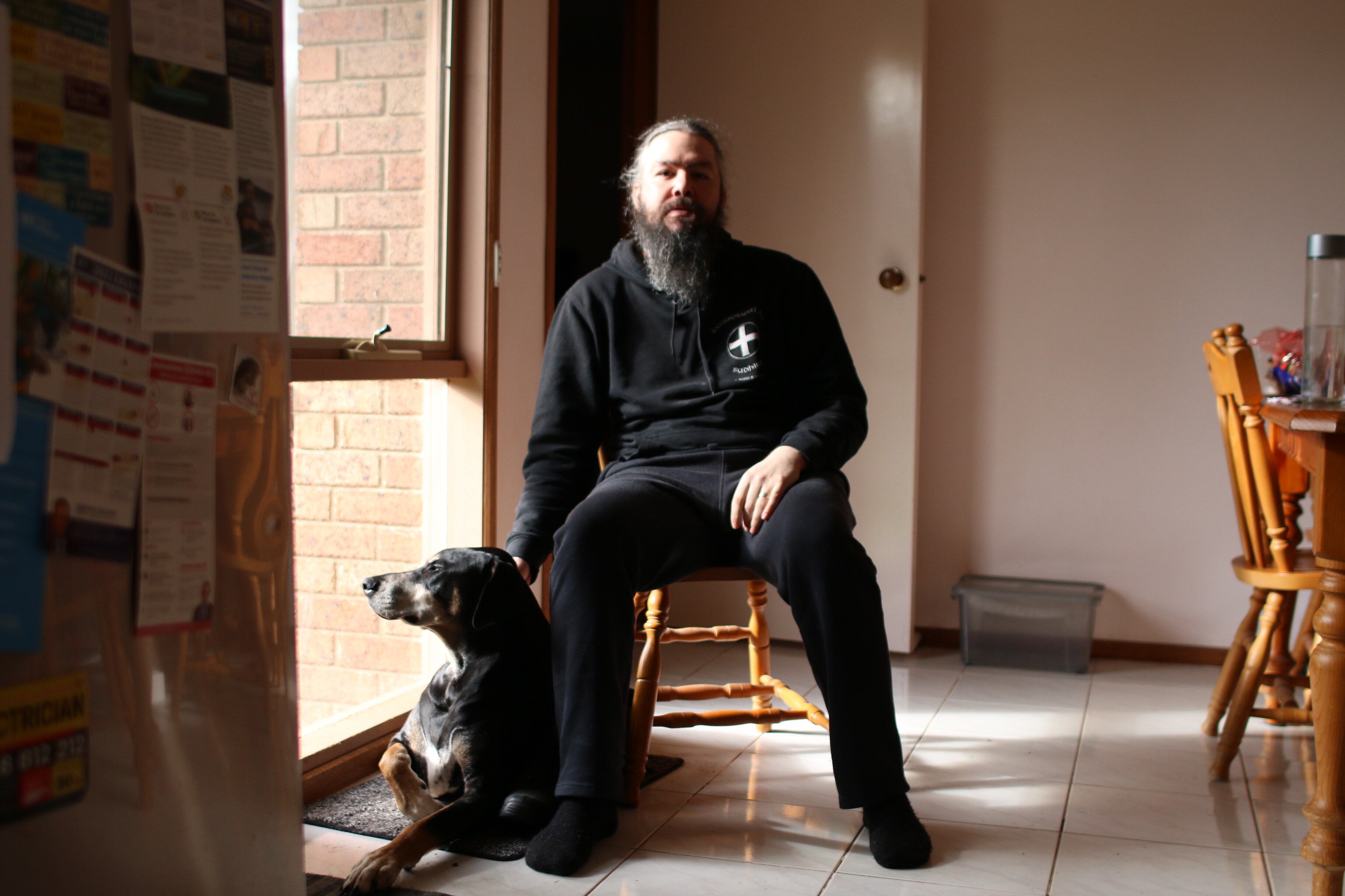 A man with long hair and a beard sitting in a kitchen with a dog