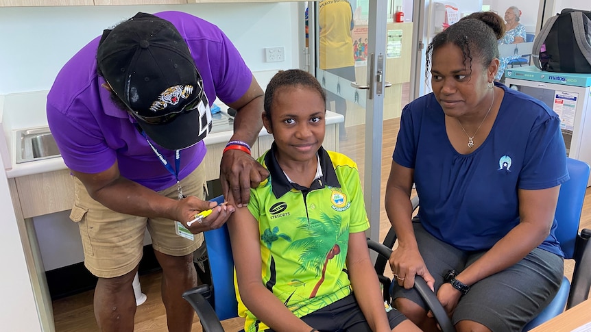 A doctor gives a girl a needle in her right arm as her mother watches.