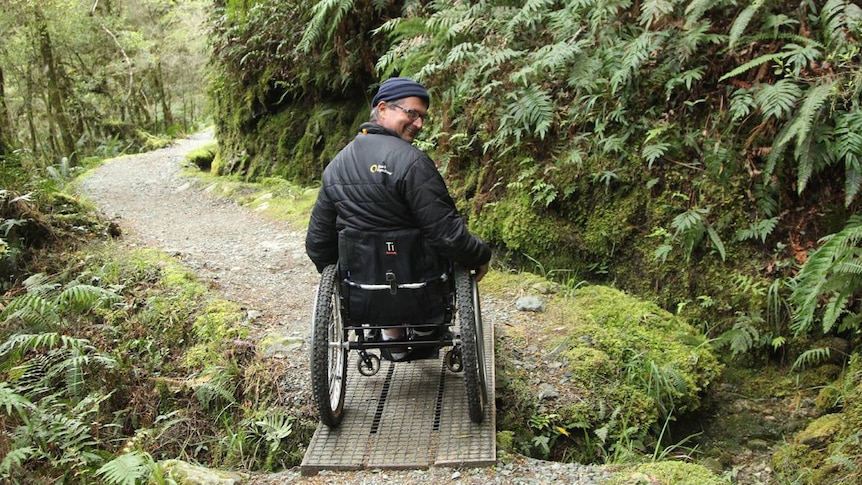 A white, middle-aged man in a wheelchair crossing a ramp in a lush forest. 