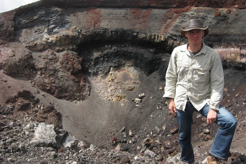 A man in a hat, jeans and a light shirt standing in front of a volcanic crater.