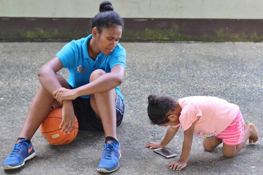 Tiyana sits with a basketball between her legs watching her daughter play with a mobile phone.