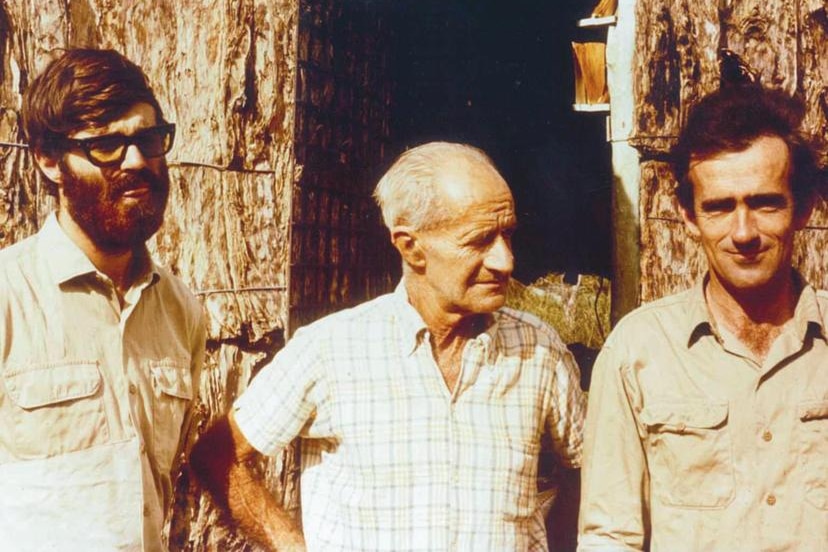 Sepia image of three white men  in front of bark hut