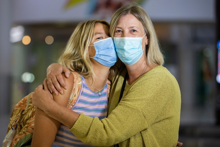 A mother and daughter stand hugging wearing face masks and posing for a photo inside Perth Airport.