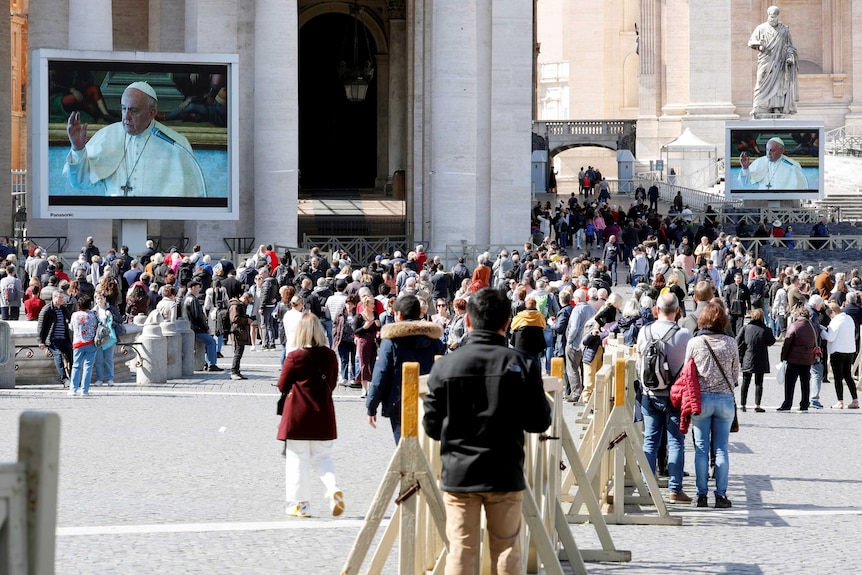 A crowd of people in an old square watch at giant TV screen showing the image of the Pope.