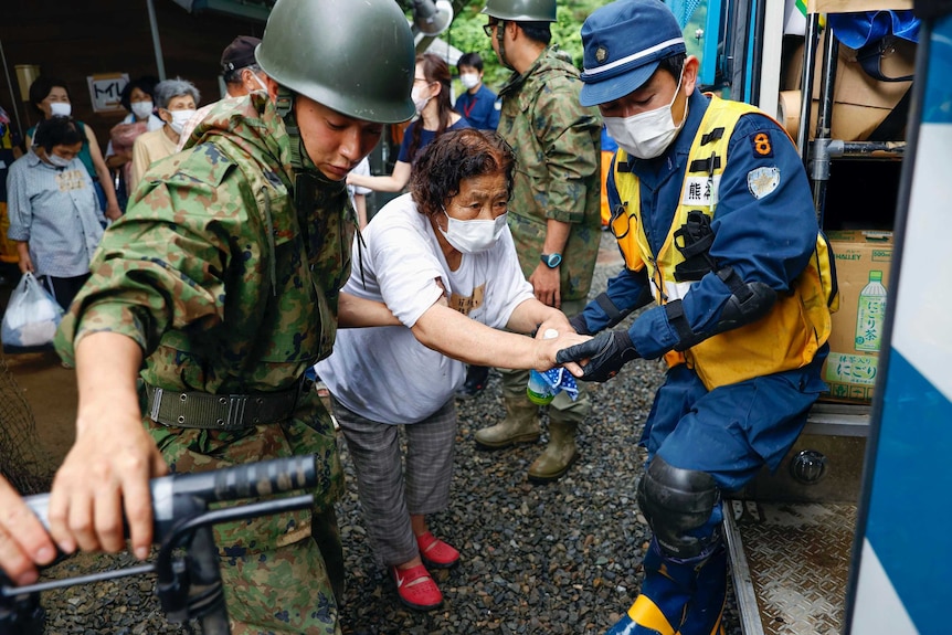 An old woman in a face mask is helped by two male emergency workers to walk from shelter into van.