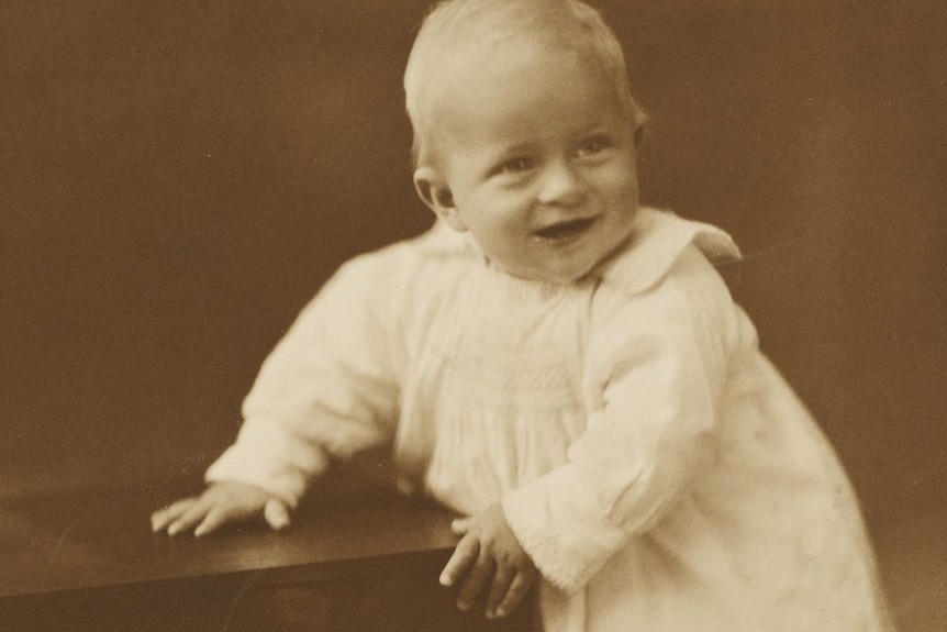 A sepia photograph of infant Prince Philip in a white dress, standing and holding onto a small table.