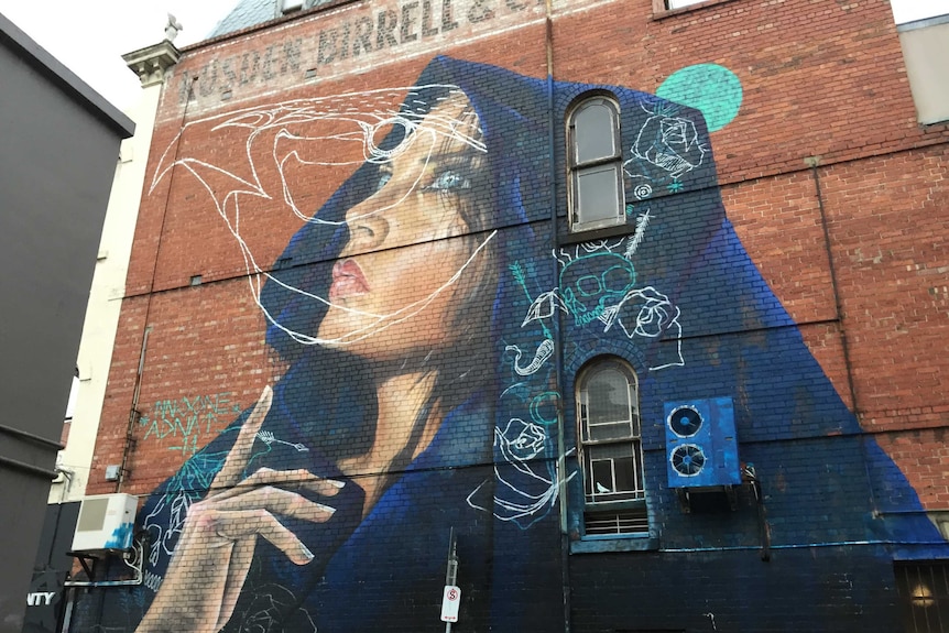 A street art mural on a two-storey wall in Fitzroy depicts a woman's face with a dark blue cloak on her head.