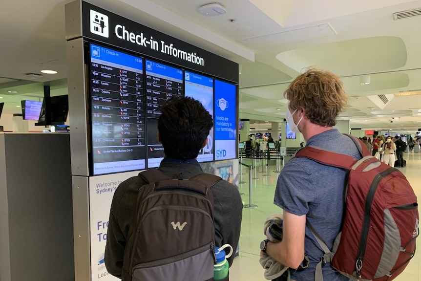 two travellers standing in front of a check in board at the airport