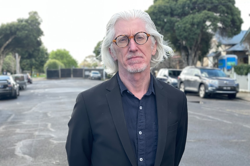 A man with long white hair and glasses in a carpark