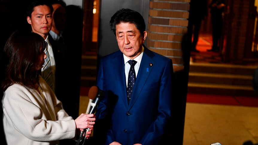 You see Shinzo Abe in a bright blue suit speaking to journalists holding a number of recording devices.