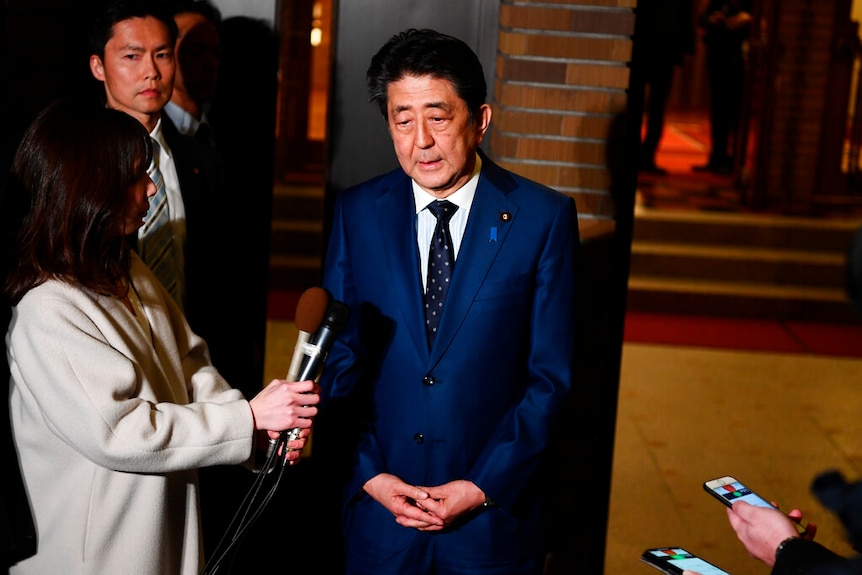 You see Shinzo Abe in a bright blue suit speaking to journalists holding a number of recording devices.