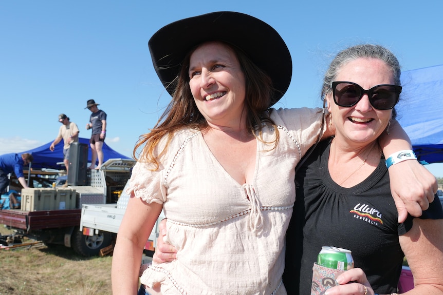 Two women smile, with one holding a can of beer. Two men stand on the back of a ute in the background.
