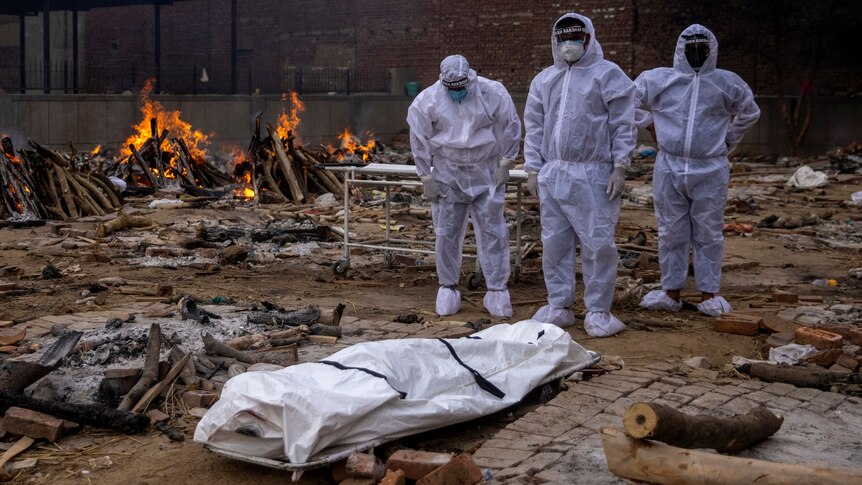 Three men in protective suits stand next to a body covered on the floor as fire burns behind them.