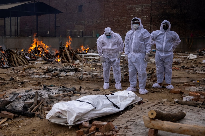 Three men in protective suits stand next to a body covered on the floor as fire burns behind them.