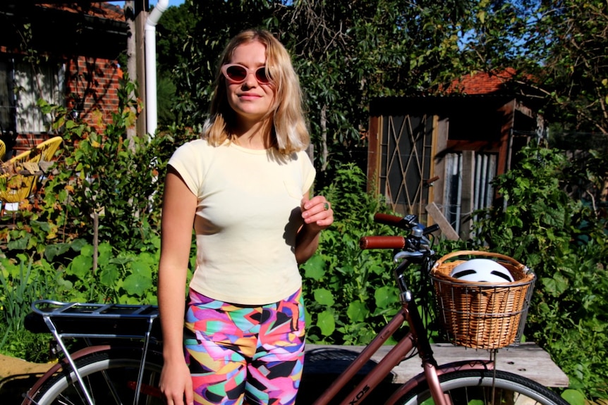 A girl with blonde hair and a pale yellow t-shirt wearing sunglasses stands in front of a bicycle.