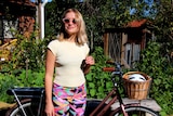 A girl with blonde hair and a pale yellow t-shirt wearing sunglasses stands in front of a bicycle.
