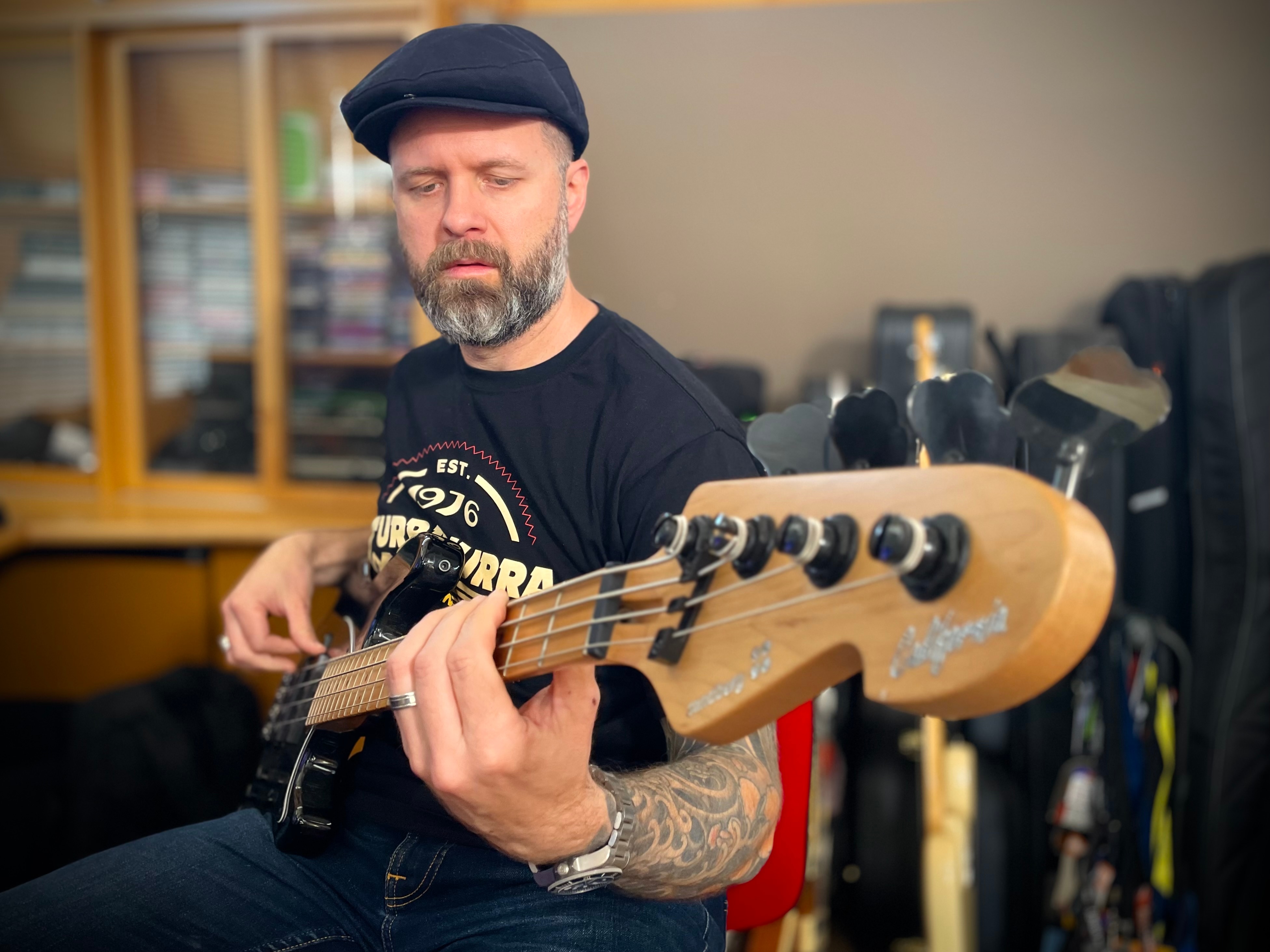 Sam Brewster plays a black bass guitar. He wears a black hat and t-shirt. His left arm has a tattoo sleeve and a watch.