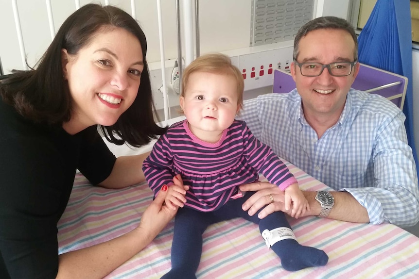 A smiling man and a woman hold on to a toddler sitting on a bed, staring inquisitively at the camera.