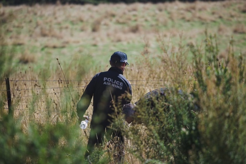 Two police officers inspect a barbedwire fence behind long grass.