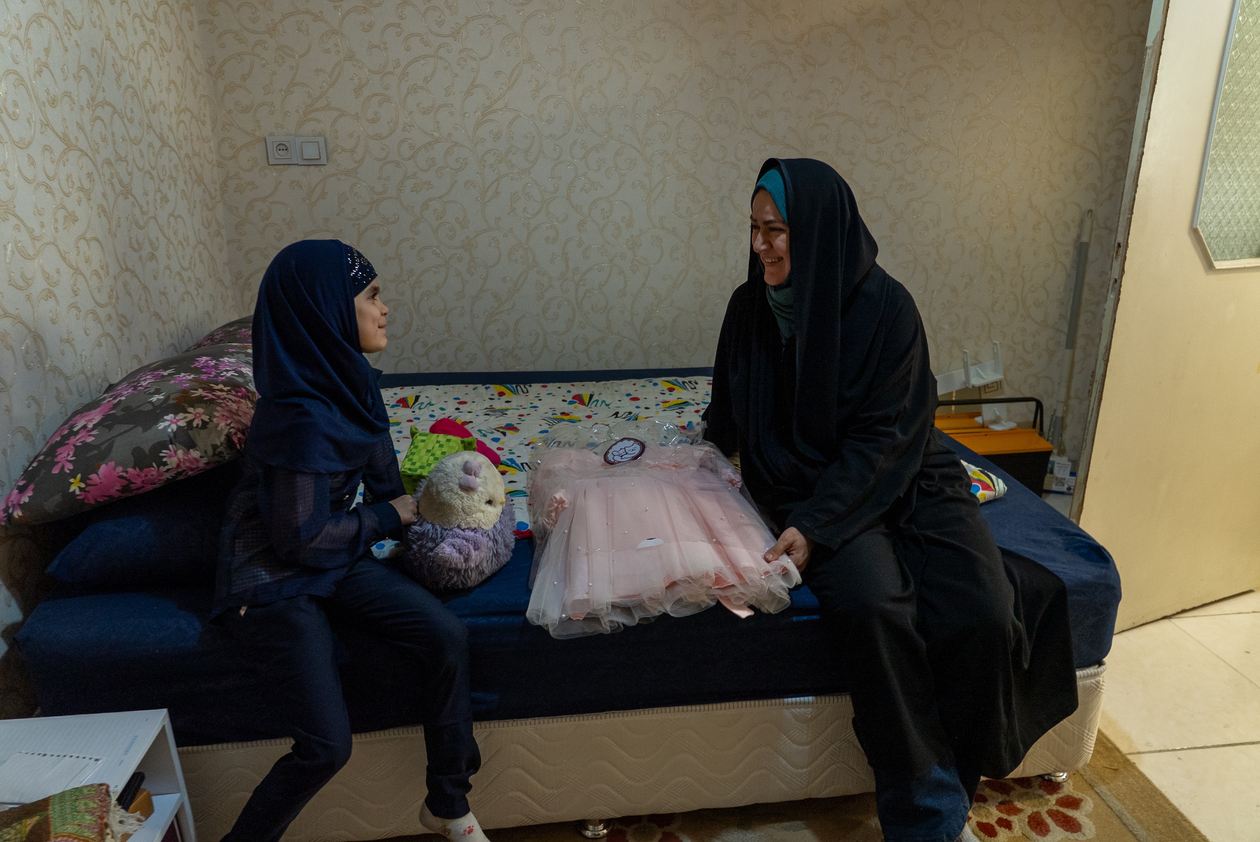 A woman and her nine-year-old daughter sit on a bed. A pink dress lies on the bed between them.