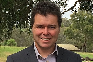 A man in his 30s smiles at the camera. His hair is black and eyes are brown. It is green behind him.