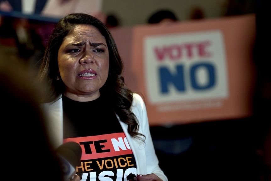 A woman with long brown hair in a white blazer standing in front of a Vote No poster.