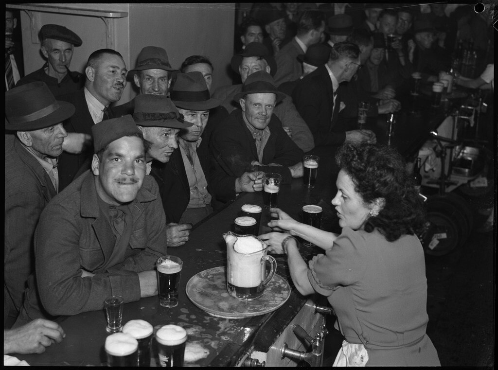 A 1940s black-and-white photo of many men cramming at a bar as one woman serves them beers