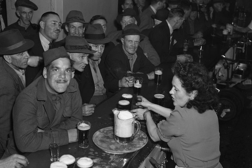 A 1940s black-and-white photo of many men cramming at a bar as one woman serves them beers