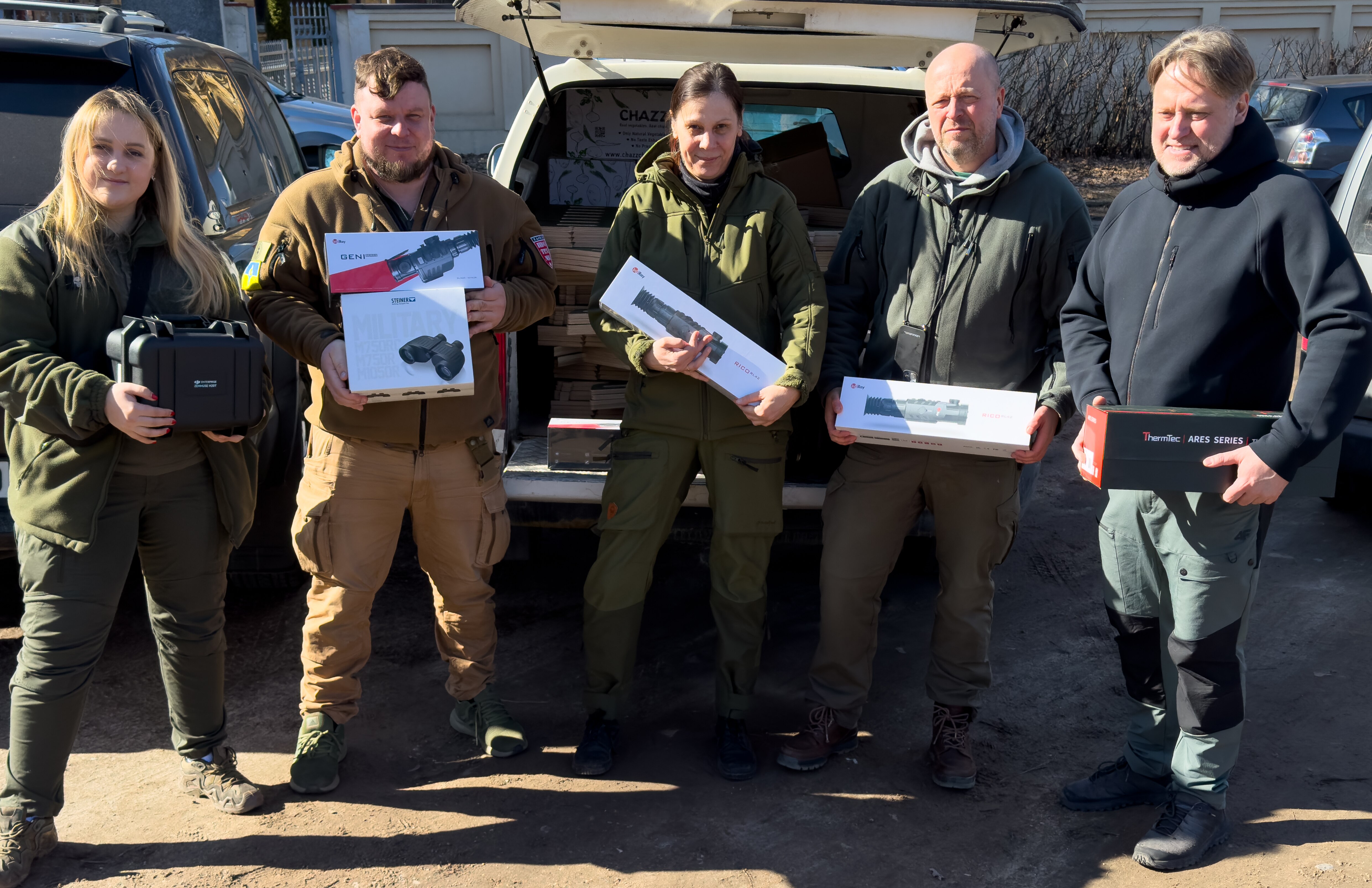 Five people stand by an open car boot, each holding a box or two containing some form of surveillance tech