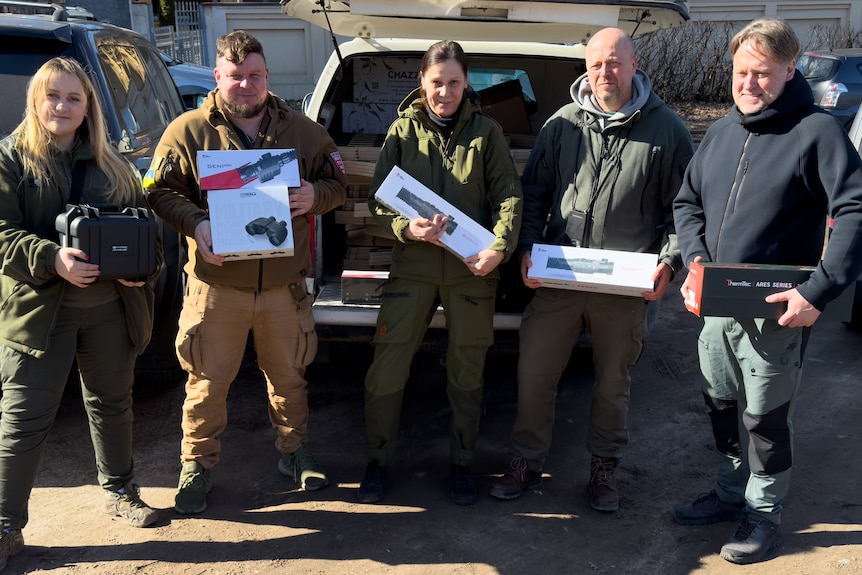 Five people stand by an open car boot, each holding a box or two containing some form of surveillance tech