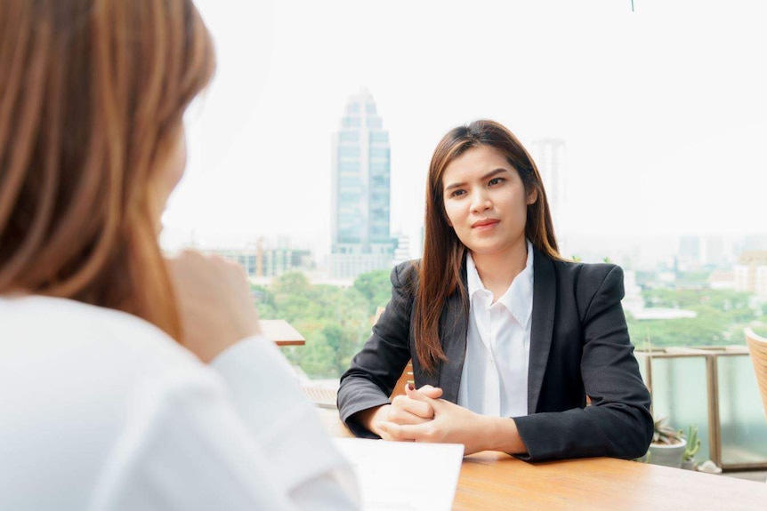 In an office, a young woman in a business suit sits at a desk facing another woman. In the background is a cityscape.