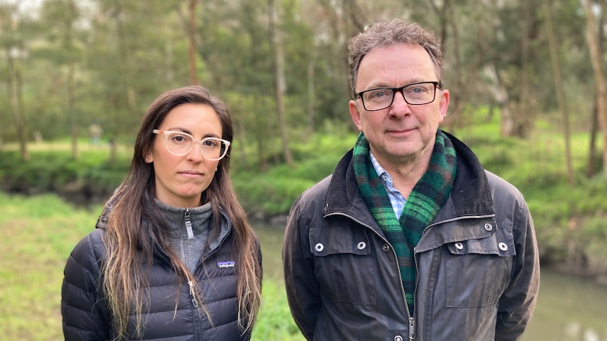 a man and a woman standing side by side with a creek behind them. They both wear glasses and jackets.