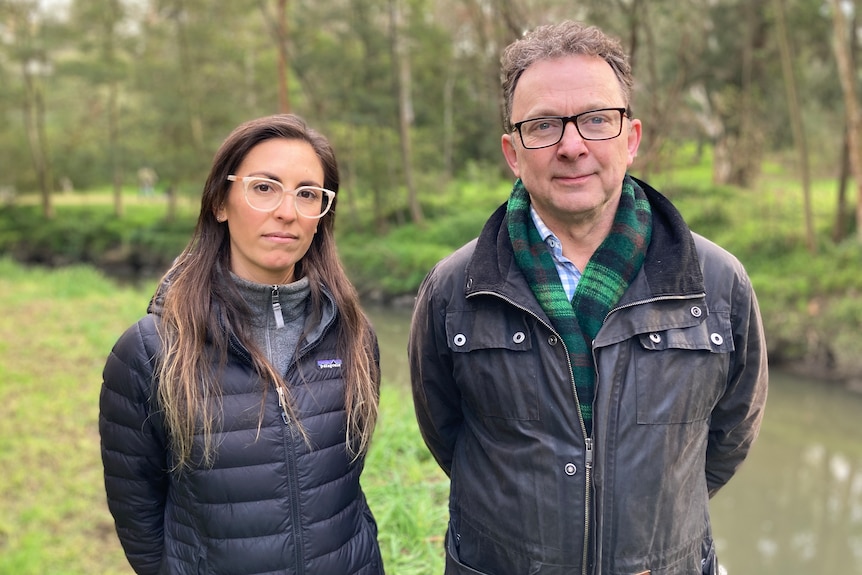 a man and a woman standing side by side with a creek behind them. They both wear glasses and jackets.