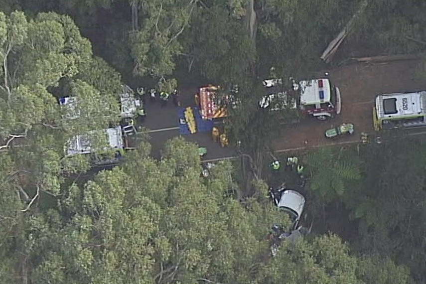 An aerial shot of a road surrounded by trees with numerous emergency services vehicles and a car off the side.