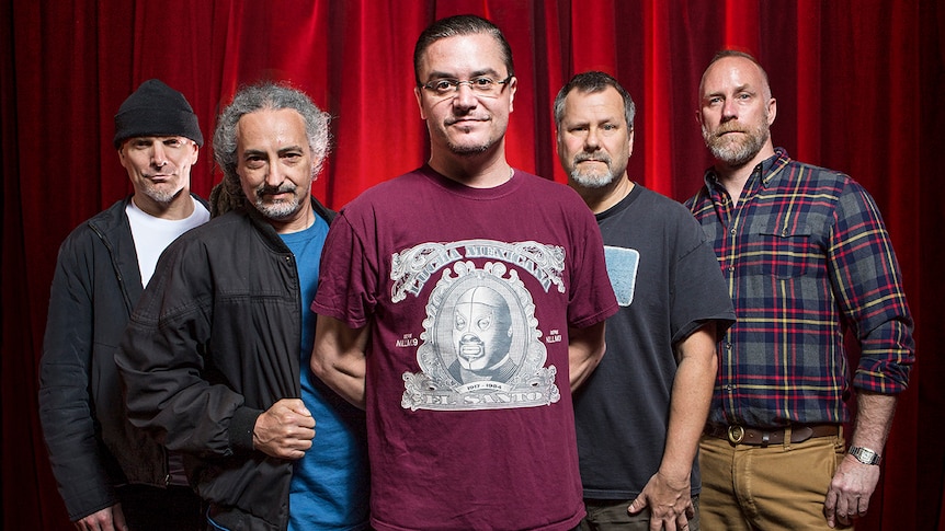 Five members of Faith No More assemble in front of a red curtain