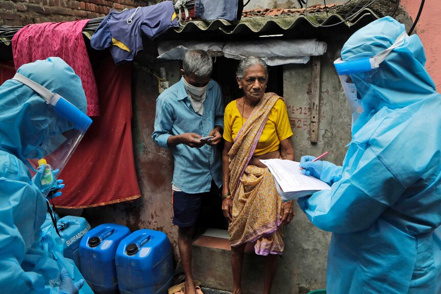 A woman in a saree and a man stand in front of a shanty as health workers in PPE write something on a form.