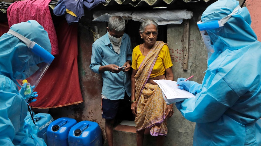 A woman in a saree and a man stand in front of a shanty as health workers in PPE write something on a form.