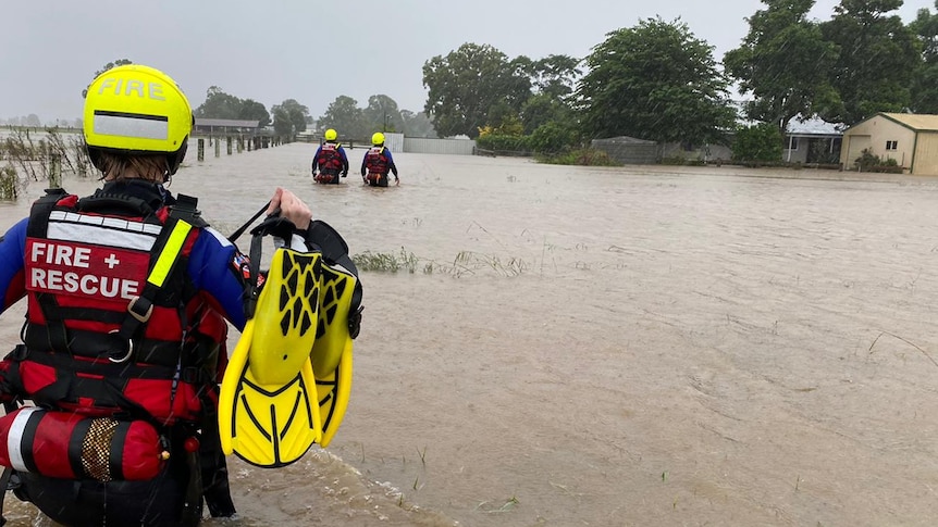 Three people wearing wet suits and helmets wade through waist deep floodwaters.