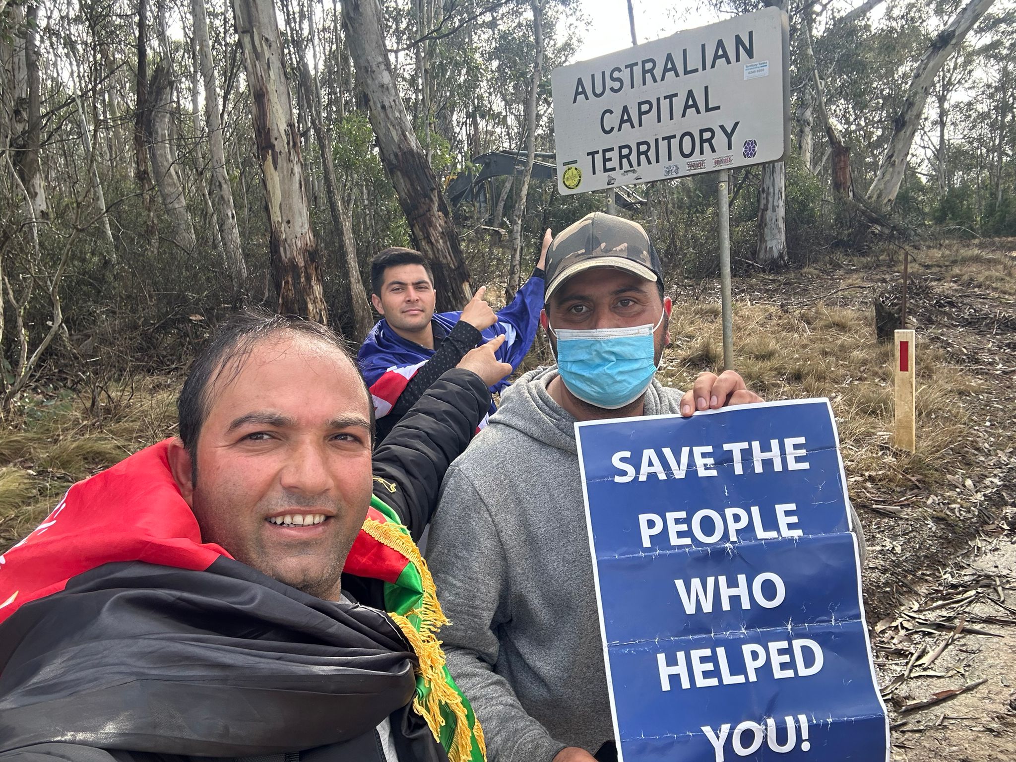 Three Afghan Australian men point to a sign reading 'Australian Capital Territory'. Two wear Afghan and Australian flags. 