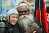 A white woman with short hair next to an Aboriginal man with a grey bear and hair, his fist clenched and a holding a flag.