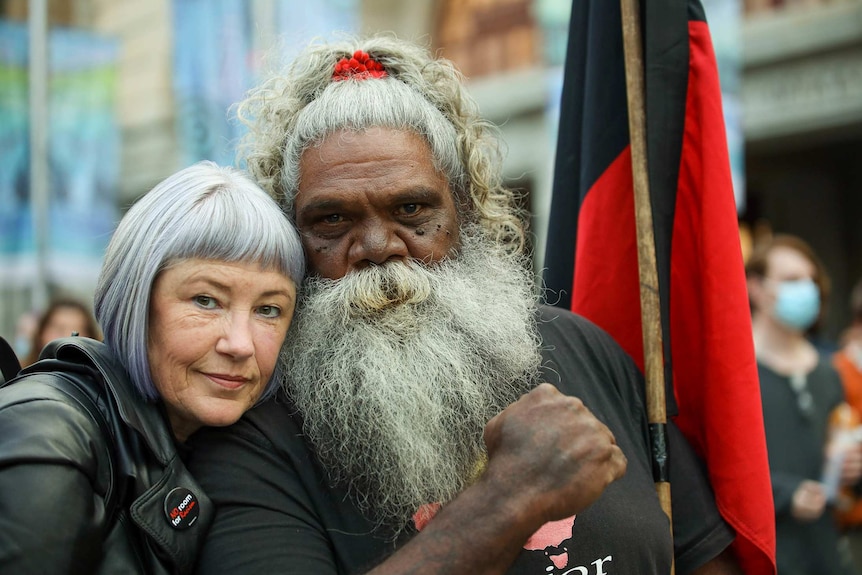 A white woman with short hair next to an Aboriginal man with a grey bear and hair, his fist clenched and a holding a flag.