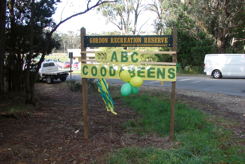 A green sign outside a country football oval with added poster that reads ABC Coodabeens in green and gold lettering
