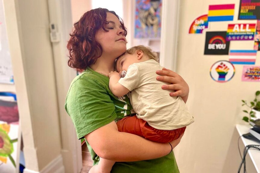 A white person with purple hair wears a green shirt and smiles holding a baby. The pair are content and happy together.