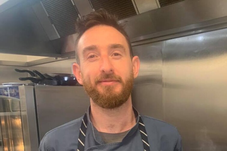 A white man with brown short hair and a beard stands in front of a deep fryer wearing a chef's apron.