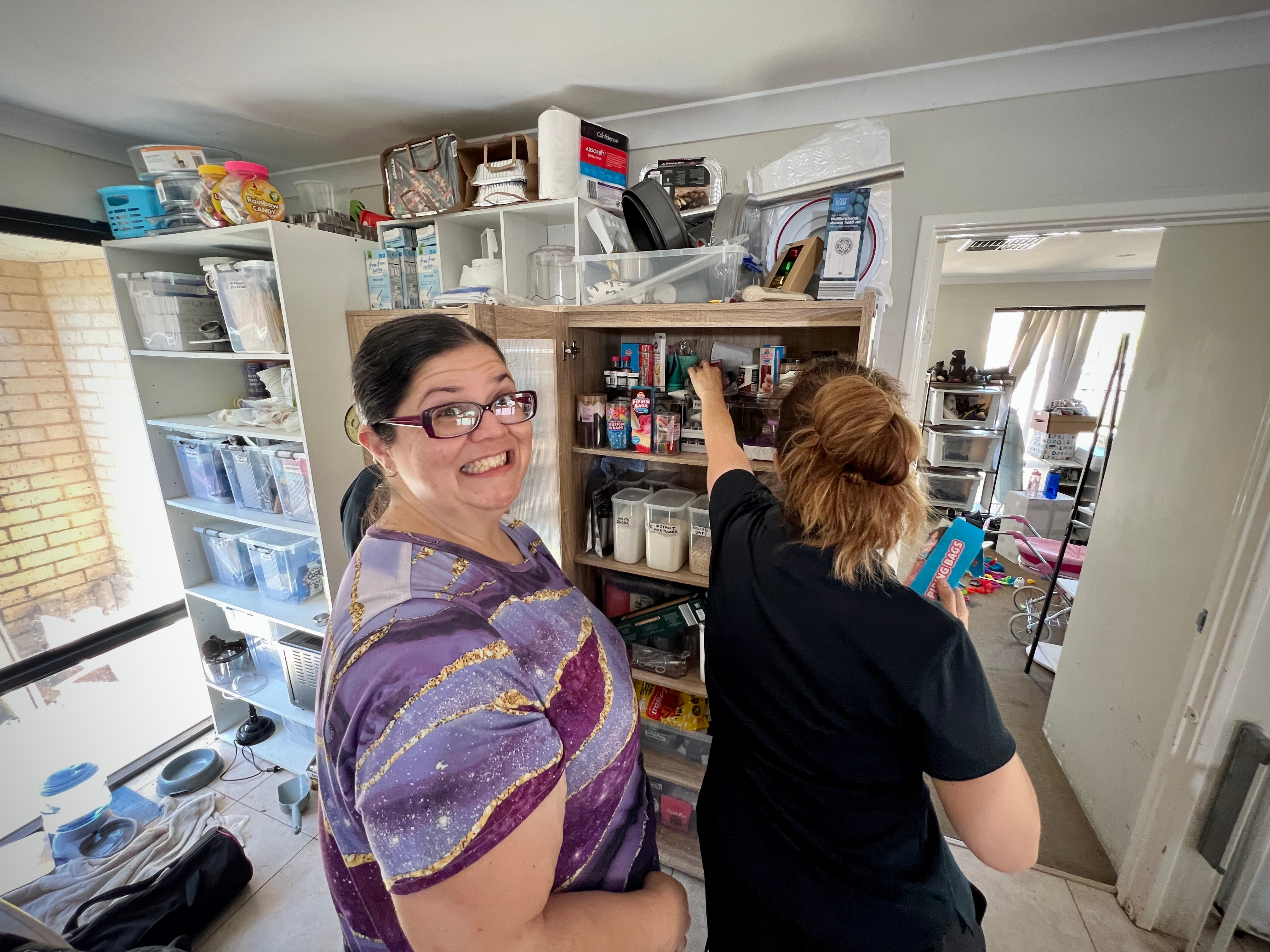Meran wears a purple shirt and looks at the camera with a humorous but apprehensive expression as a lady orders her pantry
