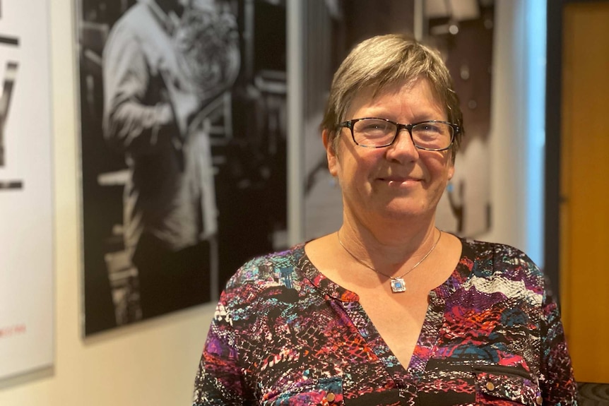 A woman wearing glasses smiles at the camera standing in front of black and white photos of instruments being played.