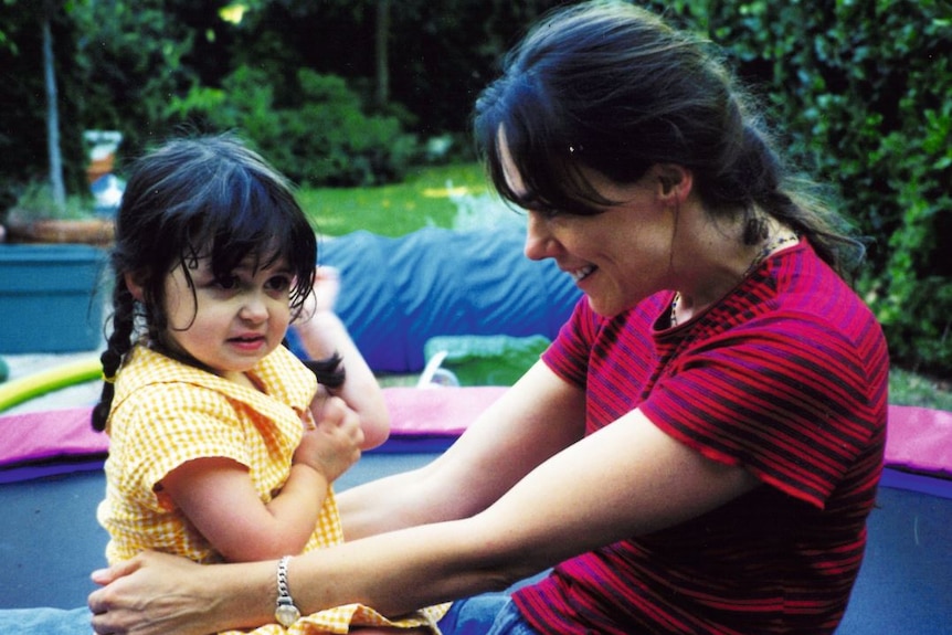 A woman and her little girl, who has pigtails in her hair, sit on a trampoline