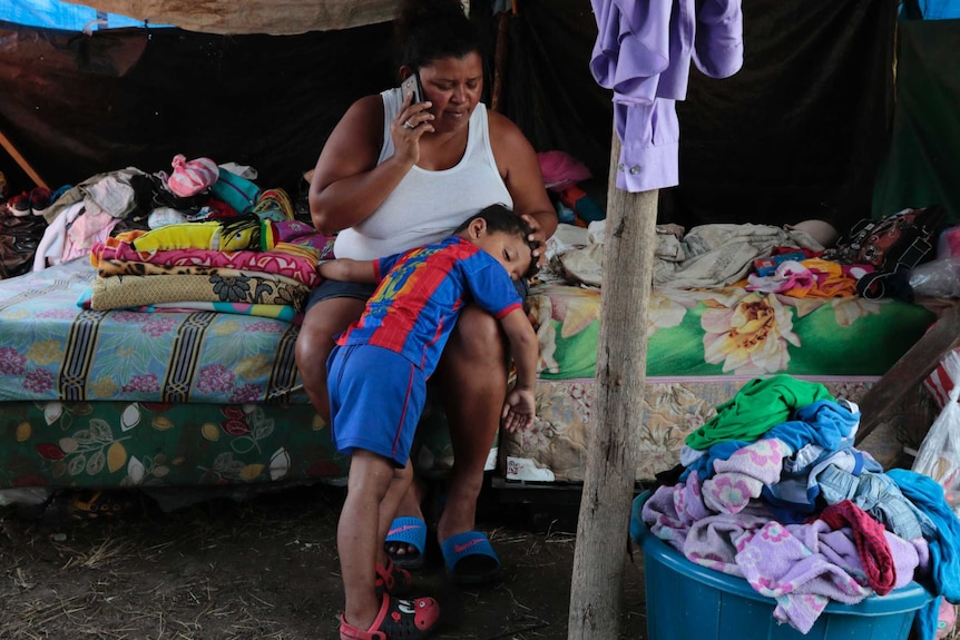 A woman sitting on a mattress under a tarp speaks on the phone as a small boy lays in her lap.