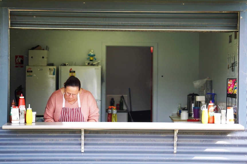 A woman in a striped apron stands behind a canteen counter lined with tomato sauce and mustard bottles.